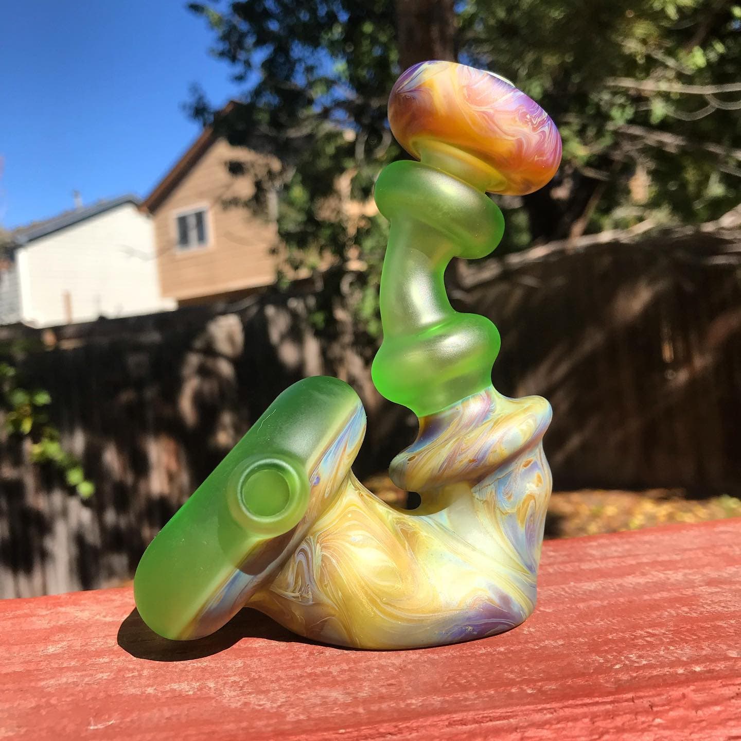 A small water piece with frosted green sections and iridescent swirl glass, sitting on a deck rail.