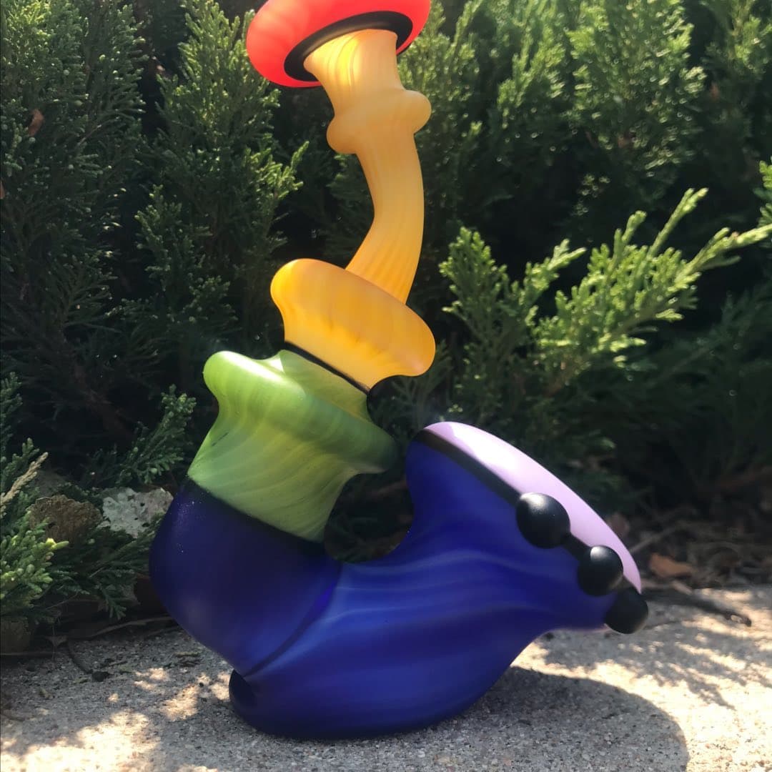 A sculptural multi-color functional pipe sitting on concrete in front of a hedge.