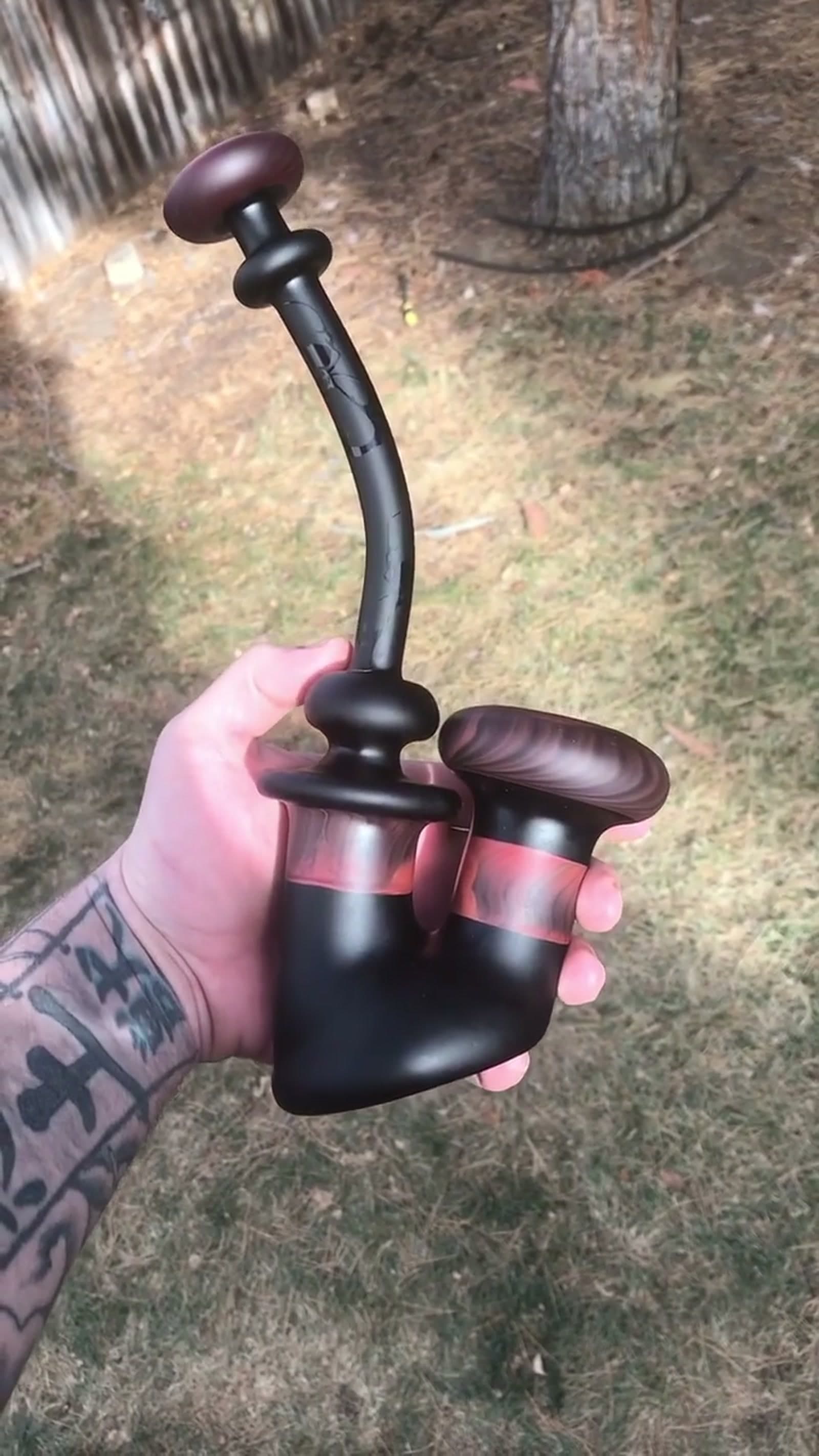 A U-shaped bubbler held in a tattooed hand, matte black with woodgrain-colored glass accents.